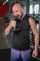 Fototapeta na wymiar bald man bites his teeth into an iron dumbbell in the gym. Sportsman with a dumbbell in mouth