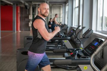 Fototapeta na wymiar Canadian Fit Muscle bald Man With Headphones Running on Treadmill in Gym.