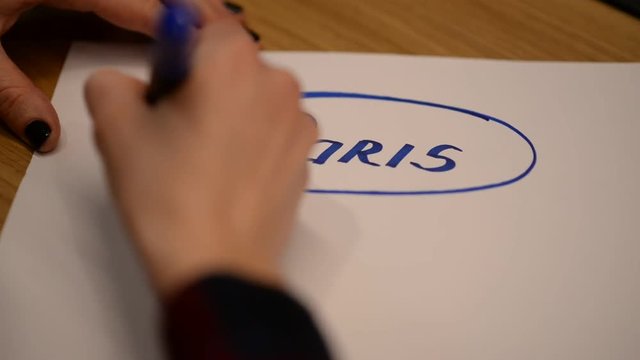 The hand of a young girl encircles the inscription Paris with a blue marker.