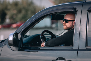 bald and bearded man in glasses with a clock in a suit behind the wheel of a car