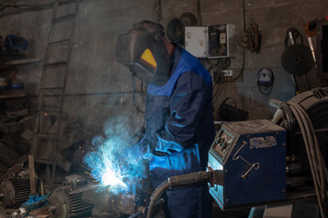 Strong professional welder is welding a metal construction in garage wearing mask, proctive glasses and blue uniform. Blue sparks are flying apart.