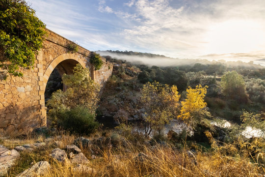 Puente medieval en paisaje otoñal con niebla y destello de sol al amanecer de un día frío.