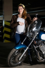 Fototapeta na wymiar Pregnant woman dressed in brown leather jacket standing with motorcycle, holding steering wheel and her large belly
