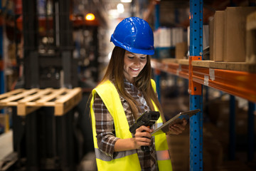 Smiling female worker holding tablet and bar code scanner checking inventory in distribution...