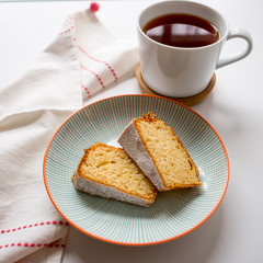 Cup of tea and two slices of sponge cake in a plate on a white table with a white and red tea towel. Top view. Squared format.