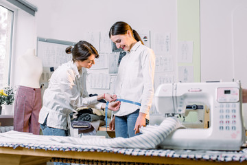 Pretty young female tailor measuring her client woman to make stylish clothes in her dressmaker studio. Garment industry, tailoring process, clothes making atelier concept