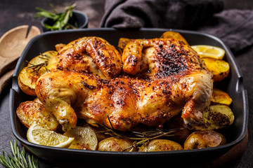 Roasted whole chicken with potatoes in a grill pan, dark background.