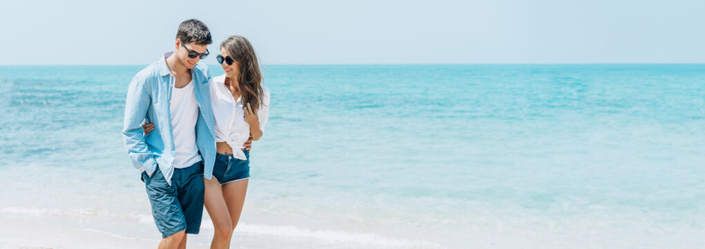 Panoramic Banner Image Of A Sweet Couple Embracing Walking On Seashore On A Beach Vacation On Coast Behind Blue Sky Or Honeymoon Trip. Wide Crop