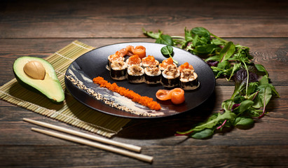 Delicious sushi rolls with caviar and sesame on black plate served on the wooden table, decorated with avocado, chopsticks and sprouts.