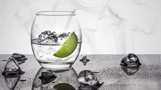 An oval glass of clear vodka, rum or gin spirit with a wedge of lime and a ice scattered black base with a smoke background