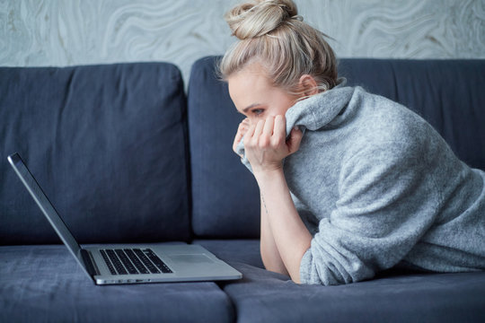 Scaried blond woman lying prone on sofa and looking on laptop computer screen