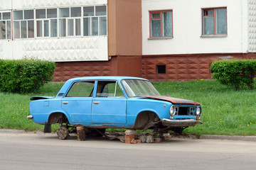 Abandoned old blue disassembled car on street near house. Old car with wheels removed, vandalism. -...