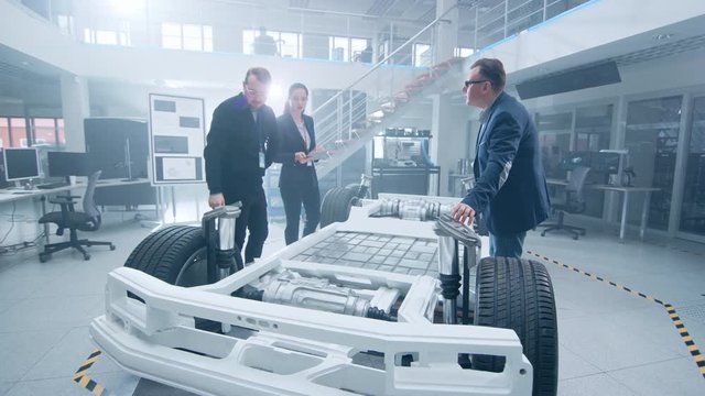 Automotive Design Engineers Talking while Working on Electric Car Chassis Prototype. In Innovation Laboratory Facility Concept Vehicle Frame Includes Wheels, Suspension, Engine and Battery.