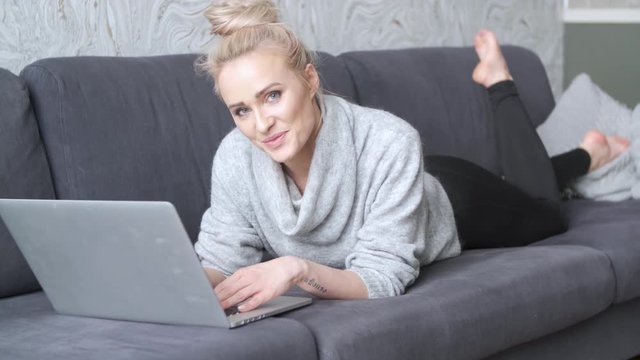 Happy blond woman lying prone on sofa and working on laptop computer