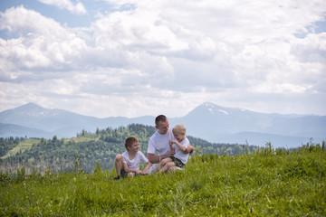 Fototapeta na wymiar Happy father with his two young sons sitting on the grass on a background of green forest, mountains and sky with clouds. Friendship concept.