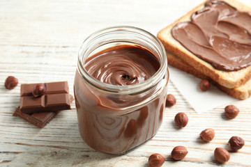 Glass jar with tasty chocolate cream, bread and hazelnuts on wooden table