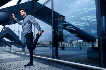 Active man doing his stretching workout next to modern building with glass wall. Urban area scenery.