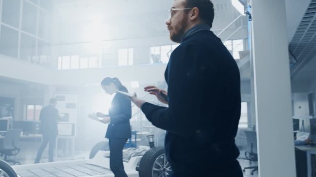 Engineer with Glasses and Beard Enters a High Tech Development Facility Holding a Tablet Computer. He Walks Towards an Electric Car Chassis Prototype with Colleagues Working on it.