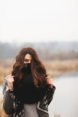 A young girl poses on the shore of a lake, covering her face with a black hat