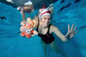 Obraz na płótnie Canvas Girl swimming instructor swims underwater and holding a toy pig - a symbol of the New year. She smiles and poses for the camera with her eyes open in Santa's hat on her head. Portrai