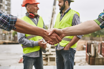 We did it! Close up photo of builders shaking hands against cheerful colleagues while working...