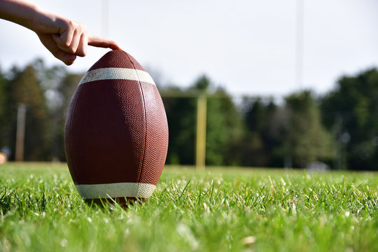Close-up photo of a football on a sunny day being held with a goal post, football field and trees in the background