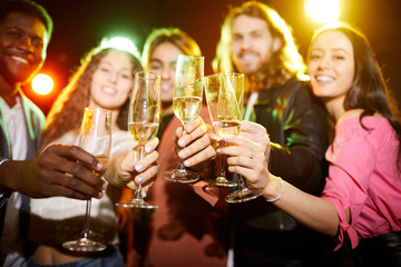 Close-up of young people in casual clothing standing in row and raising flutes while drinking champagne at party in nightclub