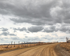 Fototapeta na wymiar view up a brown dirt road with decaying barbed wire fence and rusty gate curving under cloudy gray sky