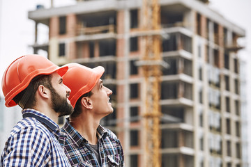 Teamwork. Two young and cheerful builders in red helmets are looking up and working at construction...