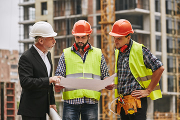 Team meeting. Main engineer in formal wear and white helmet with two young builders are discussing a construction drawing and main concept of building while standing at construction site.