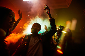 Crowd of cheerful hilarious young multiethnic people drinking alcohol and dancing in smoke at noisy party