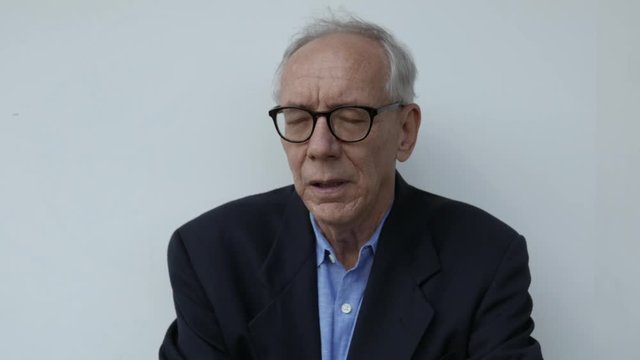 Older Man Looks Around Defeated, In Front Of A White Backdrop.