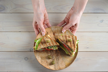 Club sandwich with ham and cheese on a wooden plate. Plate with sandwiches in two female hands. Light wooden background. View from above.