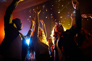 Group of carefree excited people dancing under falling confetti and waving hands in air at musical...