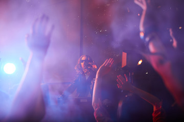 Popular handsome young dj playing record turntables and gesturing hands while interacting with crows at party in smoky nightclub