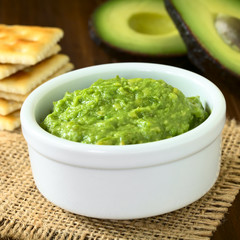 Fresh avocado cream or guacamole with soda crackers and half avocados in the back, photographed with natural light (Selective Focus, Focus in the middle of the avocado cream)