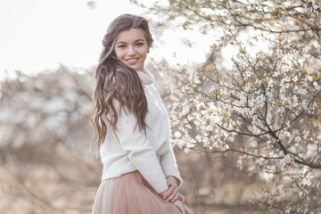 Pretty smiling teen girl are posing in garden near blossom cherry tree with white flowers.