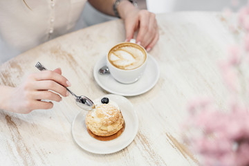 Cappuccino coffee with cup with crema in a heart wolf and cream cake dessert on a white wooden table in a cafe. Female hands with a spoon.