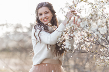 Pretty smiling teen girl are posing in garden near blossom cherry tree with white flowers. Spring time