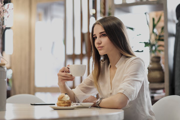 Young beautiful brown-haired girl in a white shirt is sitting at a table in a cafe by the window, drinking cappuccino coffee and dessert with whipped cream.