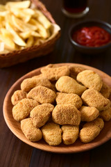 Fried breaded crispy chicken nuggets on wooden plate, French fries, ketchup and soft drink in the back, photographed on dark wood with natural light (Selective Focus, Focus one third into the image)