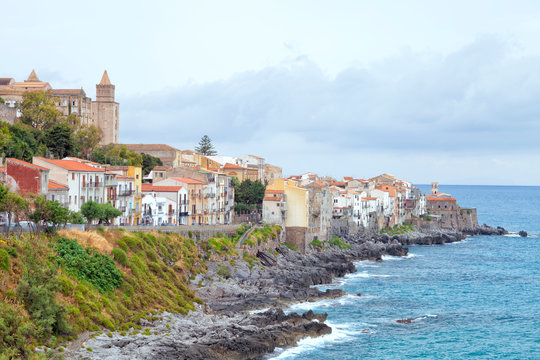 Rocky coast of Sicilian seaside resort of Cefalu with historic houses, cathedral on the hill, promenade .