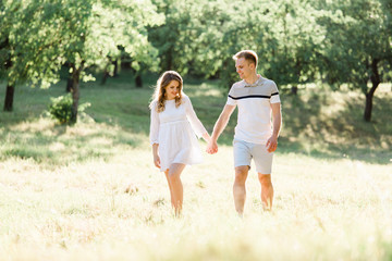 Fototapeta premium Young pretty couple in love walking in park. Handsome cheerful blonde girl in white dress hugging her boyfriend. Man and woman having fun outdoors