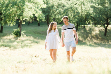 Naklejka na ściany i meble Young pretty couple in love walking in park. Handsome cheerful blonde girl in white dress hugging her boyfriend. Man and woman having fun outdoors