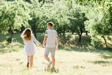 Back view of young pretty couple in love walking  in park. Handsome cheerful blonde girl in white dress hugging her boyfriend. Man and woman having fun outdoors