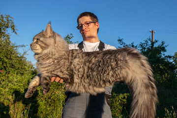 A man wearing glasses and overalls is holding a huge, gray Maine Coon cat. Cat color black silver...