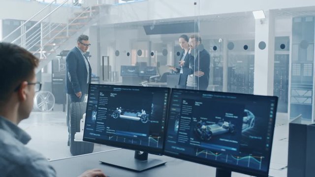 Professional Engineer Works on a Computer with a 3D Software while Colleagues Inspect Electric Car Chassis Prototype with Wheels, Batteries and Engine Standing in a High Tech Development Laboratory.