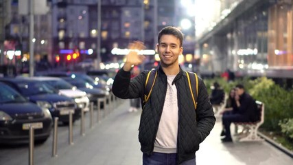 Young happy attractive smiling man standing in the evening city street and waving hand. Show goodbye or hello gesture.