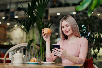 Young beautiful girl sitting in a green cafe at a table. Drinking tea with croissants, chatting, laughing and taking pictures on a smartphone.