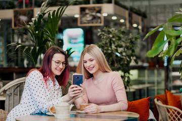 Two young beautiful girls sit in a green cafe at a table. Drink tea with croissants, chatting, laughing and taking pictures on a smartphone.